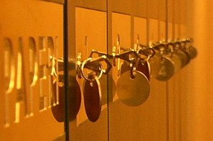 Rivissä roikkuvat avaimet, joissa on pyöreät ja soikeat avaimenperät, kiinnitettynä kultaiseen seinään, jossa lukee "PAPERI", lämpimässä valaistuksessa.