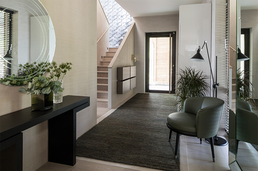 Sisääntuloaula, jossa on pyöreä peili, musta konsolipöytä, vihreä nojatuoli, matto ja portaat. Valoisa ja moderni sisustus.