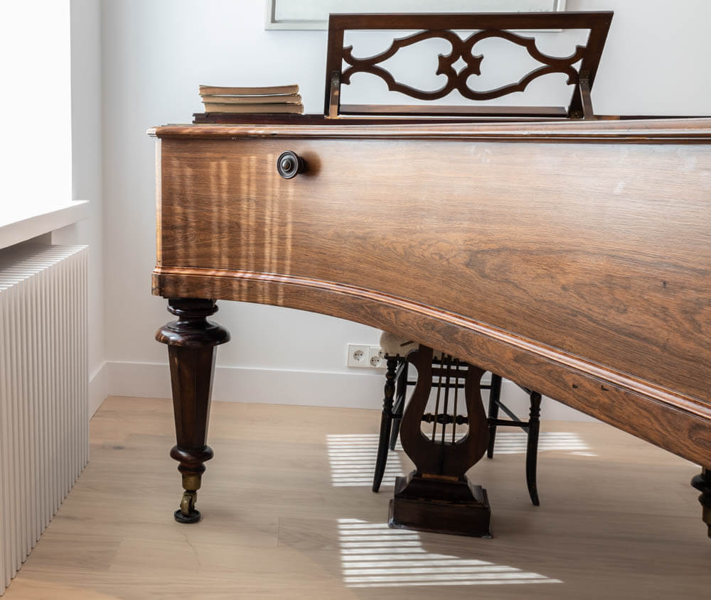 Ruskea, antiikkinen flyygeli, jossa on koristeellinen nuottiteline ja kirjapino päällä, seisoo valoisassa huoneessa puulattialla.