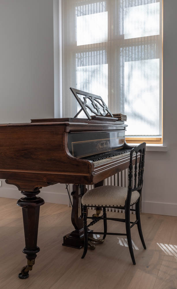 Ruskea flyygeli, musta tuoli ja valoisa ikkuna valkoisilla sälekaihtimilla valoisassa huoneessa.