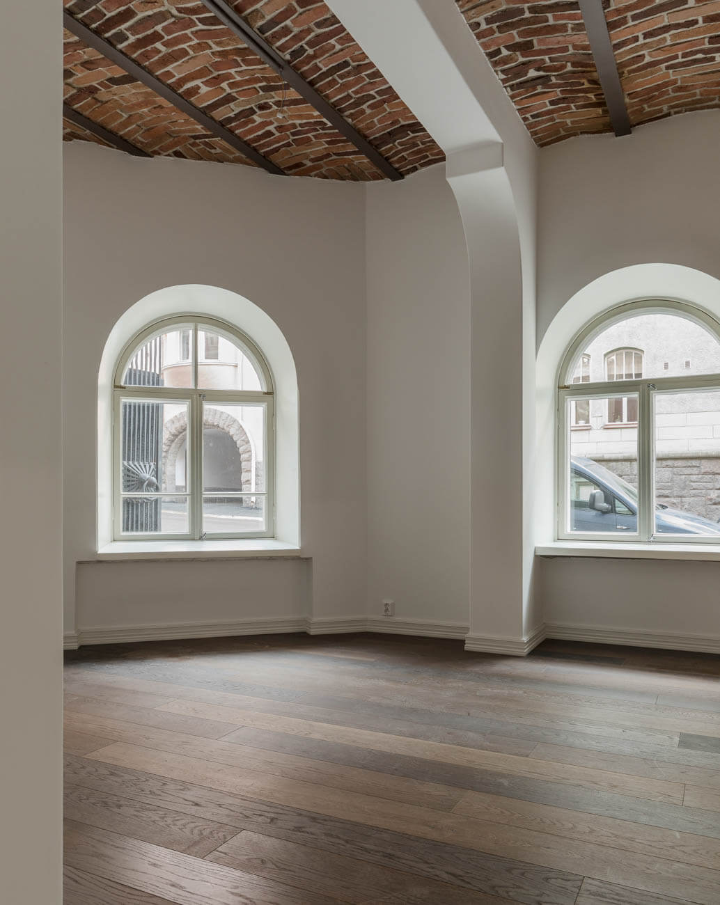 Huoneessa on tumma puulattia, valkoiset seinät ja kaksi kaari-ikkunaa. Katossa on tiilimuuraus ja metallipalkit.