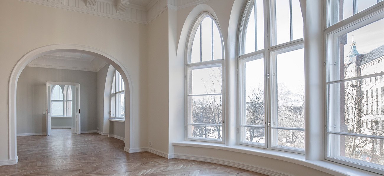 Vaaleassa huoneessa on suuret kaari-ikkunat, parkettilattia ja kaareva oviaukko, joka johtaa toiseen huoneeseen, jossa on myös ikkunoita.