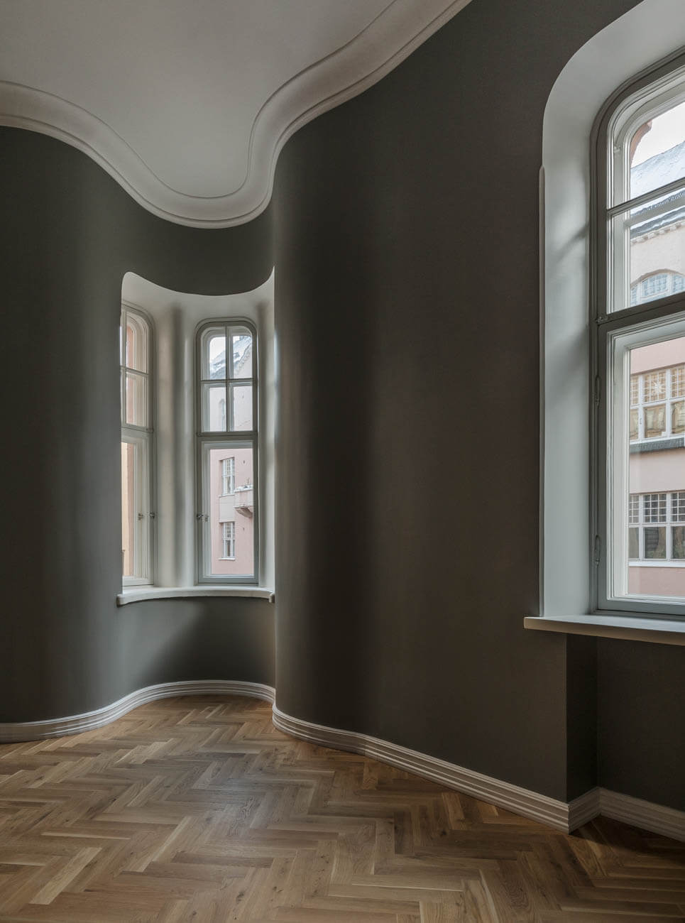 Kaareva huone, jossa on parkettilattia, kaksi ikkunaa ja harmaat seinät valkoisilla listoilla.