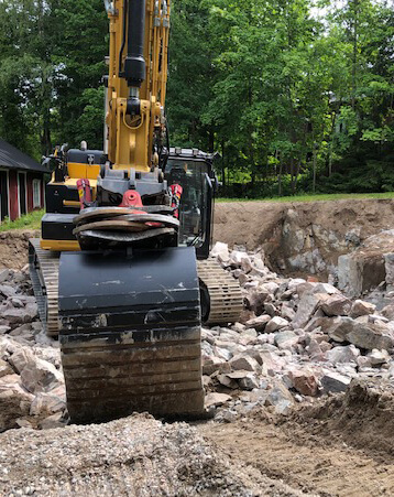 Keltainen kaivinkone työskentelee kivikkoisella alueella, jossa on paljon kiviä ja maata. Taustalla näkyy puita ja rakennus. Valoisa ja aurinkoinen sää.