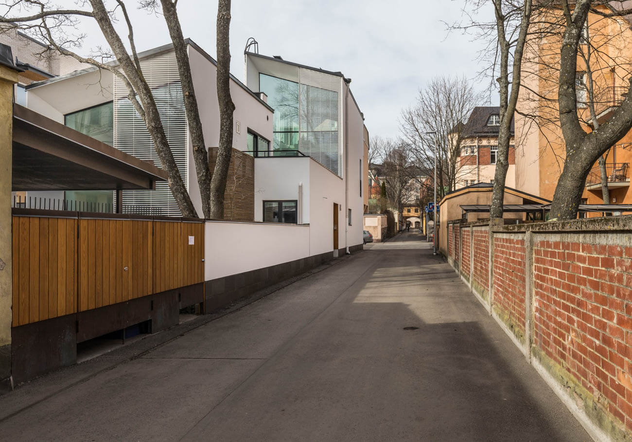 Kapea katu, jota reunustavat modernit rakennukset ja punatiilinen aita, valaistuksena pehmeä päivänvalo.