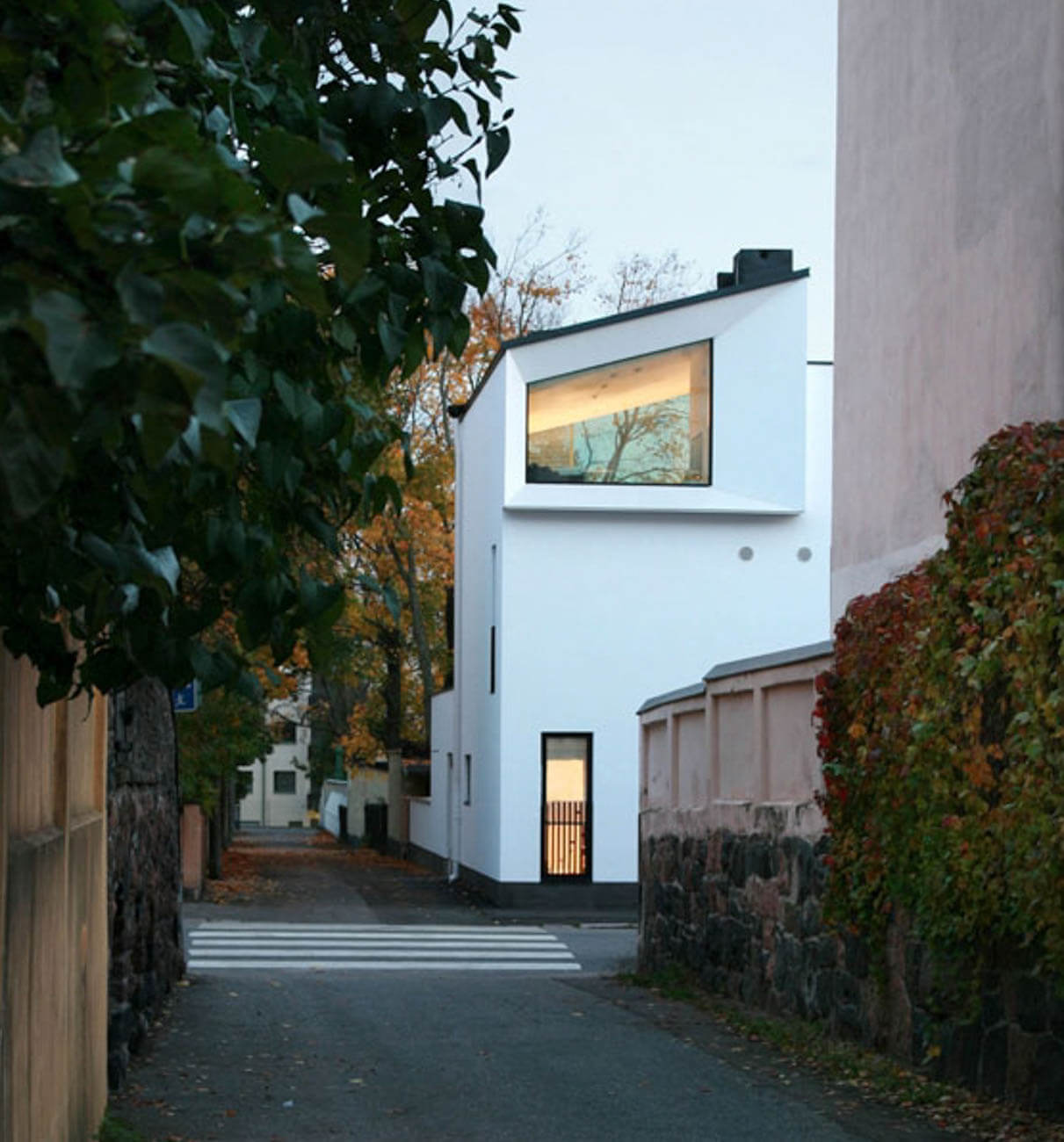 Moderni valkoinen rakennus, jossa on suuri ikkuna ja tumma ovi, sijaitsee kapealla kadulla, jota reunustavat kivimuurit ja puut.