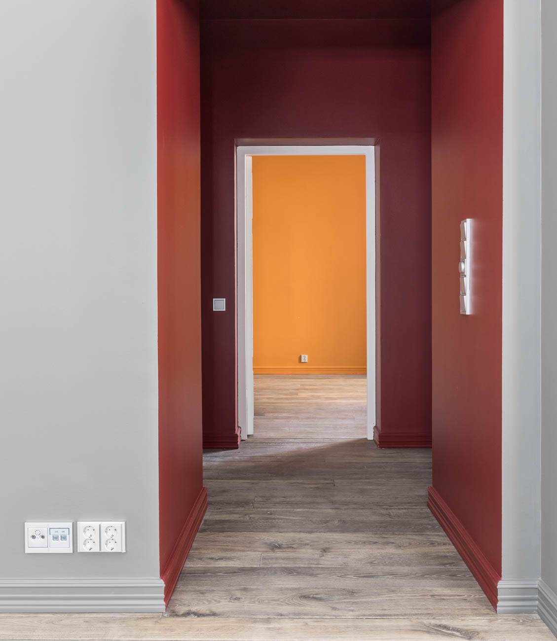 Sisätilakuva, jossa näkyy käytävä, jonka seinät ovat harmaat, punaiset ja oranssit, sekä puulattia ja valkoiset pistorasiat.