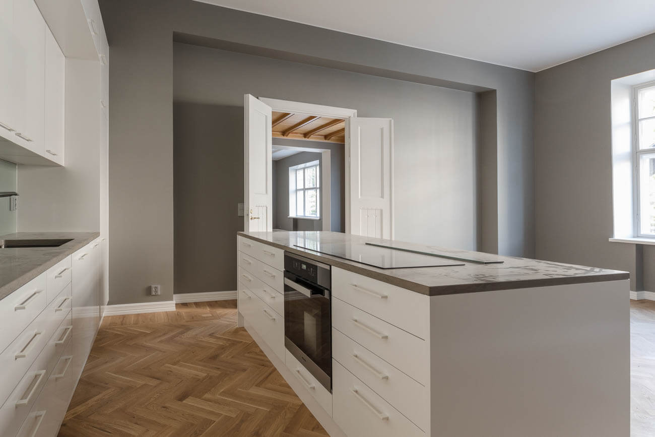 Vaaleanharmaa keittiö, jossa valkoiset kaapit, tumma työtaso ja kalanruotokuvioinen lattia. Avoimesta oviaukosta näkyy toinen huone.