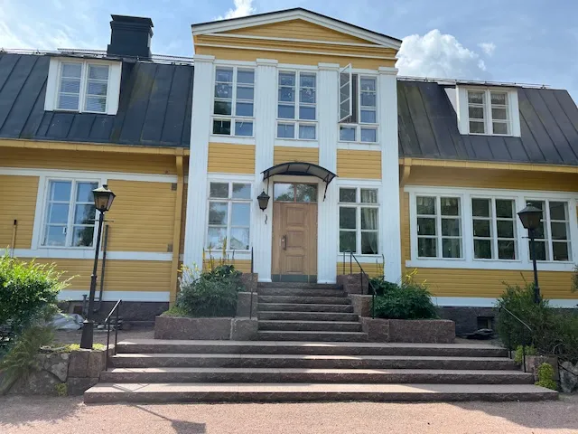 Kaksikerroksinen keltainen puutalo, jossa on valkoiset ikkunat ja pylväät, sekä kiviset portaat ja musta katto.