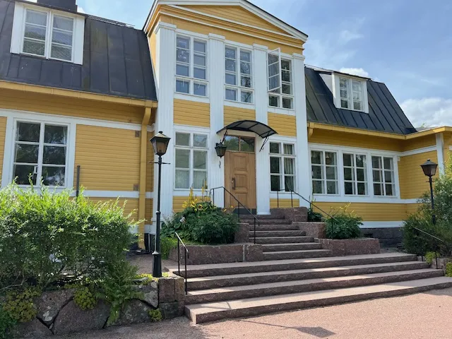 Keltaisen puutalon edessä on kiviset portaat, joita reunustavat lyhtypylväät ja istutukset. Talon katto on tumma ja ikkunat valkoiset. Aurinkoinen sää valaisee rakennuksen.