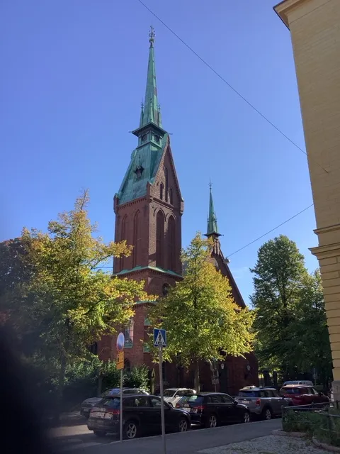 Ruskeatiilinen kirkkorakennus, jonka vihreä kuparikatto ja korkeat tornit erottuvat selkeästi sinistä taivasta vasten kaupunkialueella.