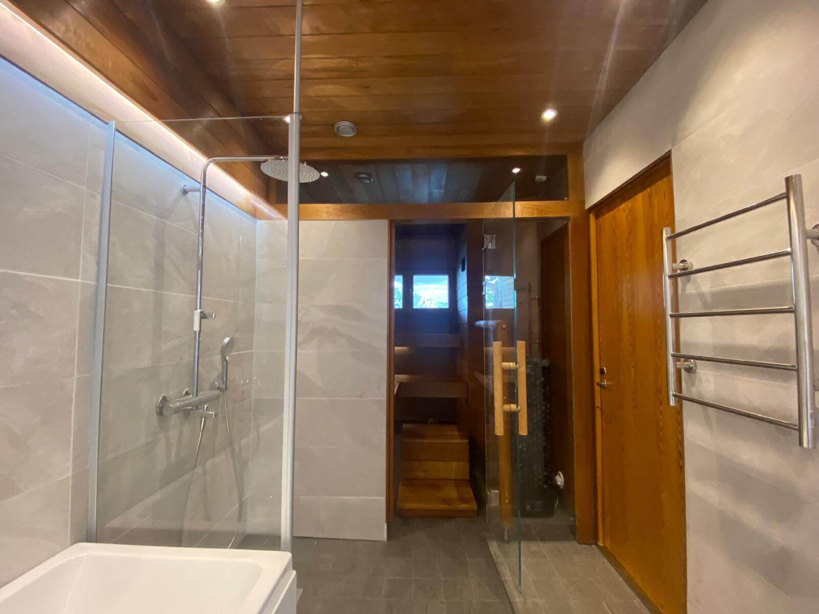 Kylpyhuoneessa on suihku, sauna ja puinen katto, valaistus on kirkas ja värit ovat pääosin harmaita ja ruskeita.