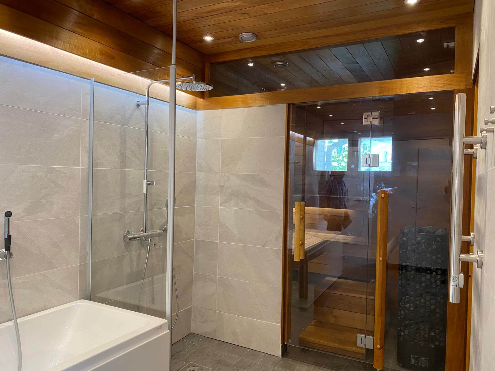 Kylpyhuoneessa on suihku, kylpyamme ja lasiovinen sauna, jossa on puupanelointi katossa ja seinissä, sekä harmaat laatat seinissä ja lattialla.