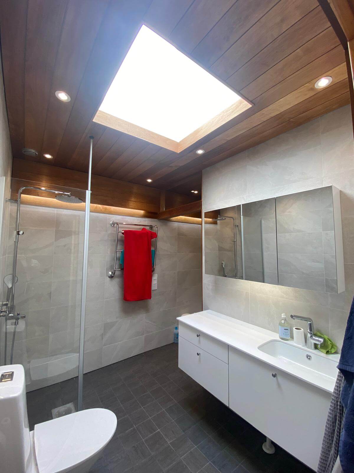 Kylpyhuoneessa on puinen kattoikkuna, harmaat laatat, valkoinen pesuallas ja punainen pyyhe.