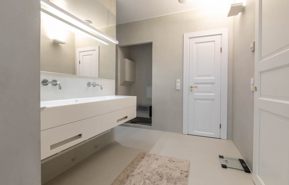 Moderni kylpyhuone, jossa valkoinen pesuallas, peili, valaistus, matto ja avoin ovi.