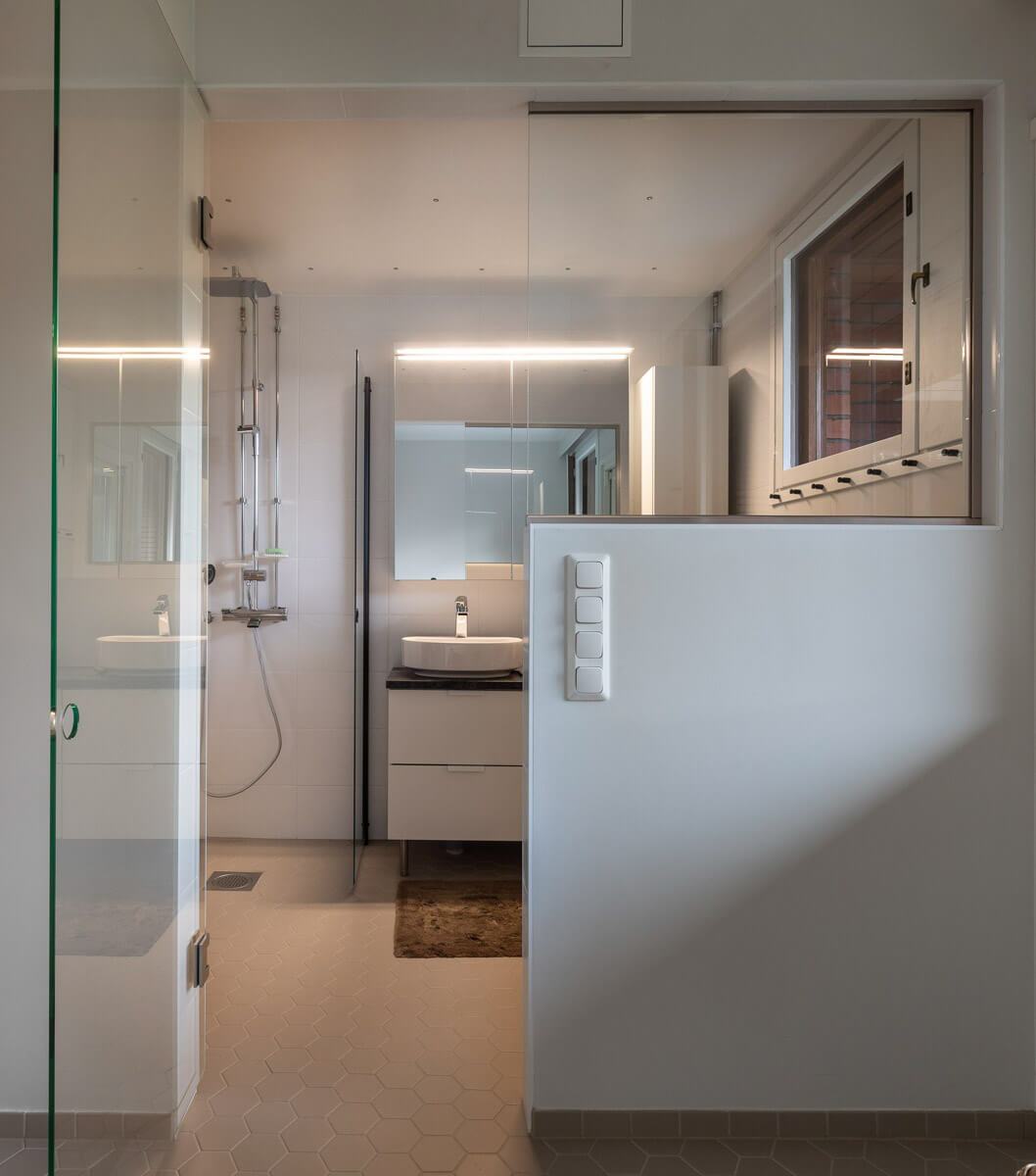 Kylpyhuoneen sisustus, jossa on lasinen suihkukaappi, pesuallas ja peili valaistuksella, sekä valkoiset seinät ja kuusikulmaiset lattialaatat.
