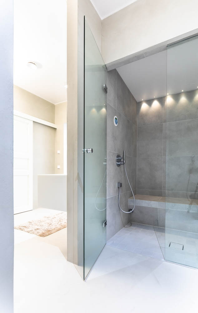 Moderni kylpyhuone lasisella suihkukaapilla, harmaalla laatoituksella ja valkoisella lattialla, jossa on beige matto näkyvissä.