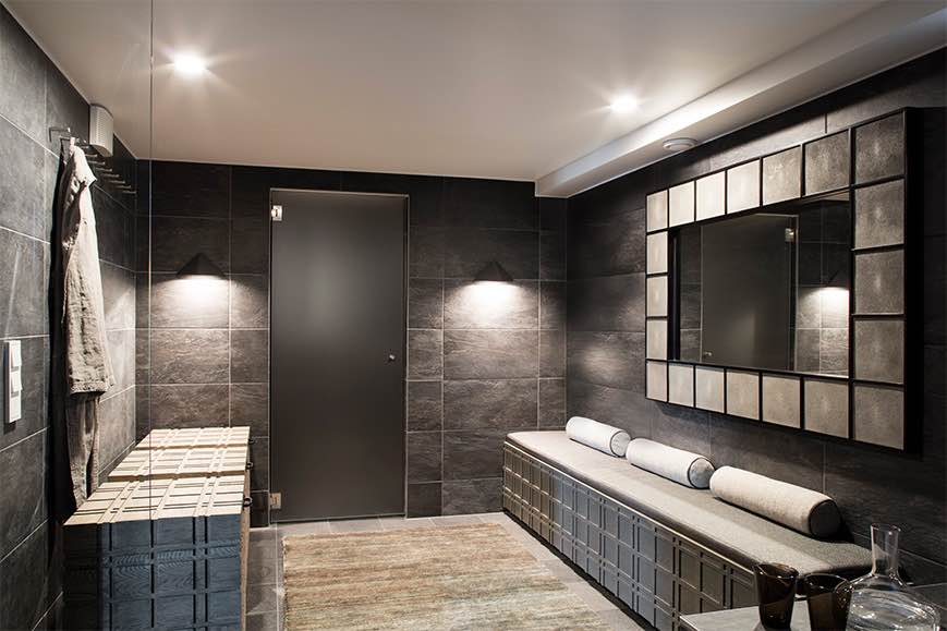 Tumma kylpyhuone, jossa on harmaa laattaseinä, peili, penkki, puinen säilytyslaatikko ja matto.