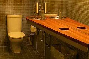 Kylpyhuoneessa on valkoinen wc-istuin, puinen pesuallas kahdella hanalla ja pieni roskakori, kaikki keltaisen ja ruskean ruudullisen seinän vieressä.