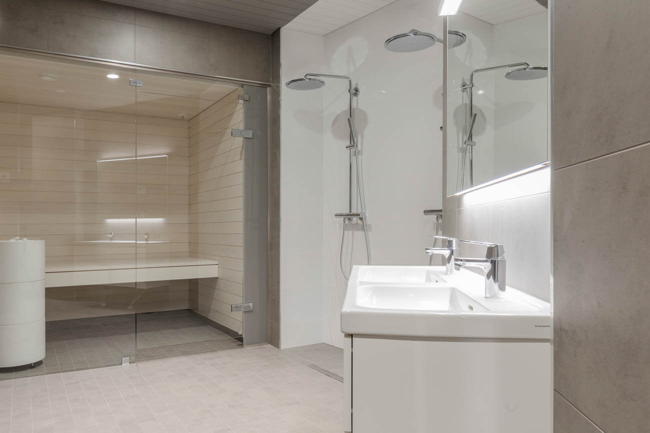 Moderni kylpyhuone, jossa on sauna, kaksi pesuallasta, suihku ja peili valaistuksella. Värit ovat vaaleita ja valaistus on kirkas.