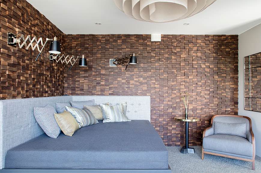 Makuuhuoneessa on puinen seinä, harmaa sänky tyynyillä, nojatuoli ja moderni kattovalaisin. Seinällä on säädettävät valaisimet.