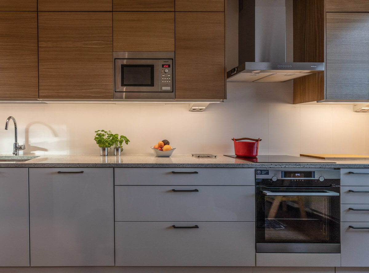 Moderni keittiö, jossa on puukaapit, harmaat alakaapit, ruostumattomasta teräksestä valmistettu mikroaaltouuni ja liesituuletin sekä punainen kattila liedellä.