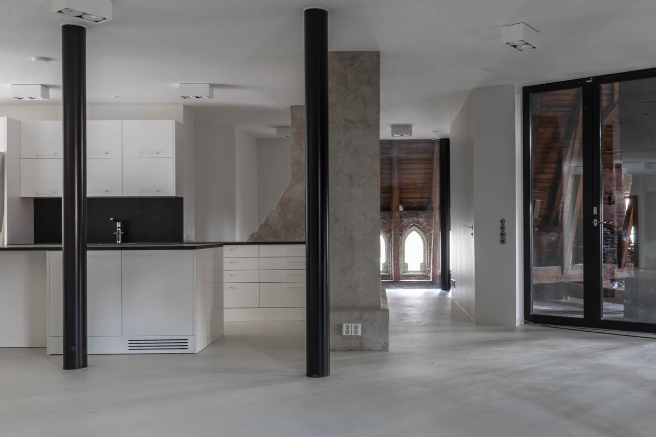 Moderni sisustus, jossa valkoiset keittiökaapit, mustat työtasot, mustat pylväät ja harmaat betonipilarit. Taustalla näkyy vanha kirkko holvikaari-ikkunoineen.