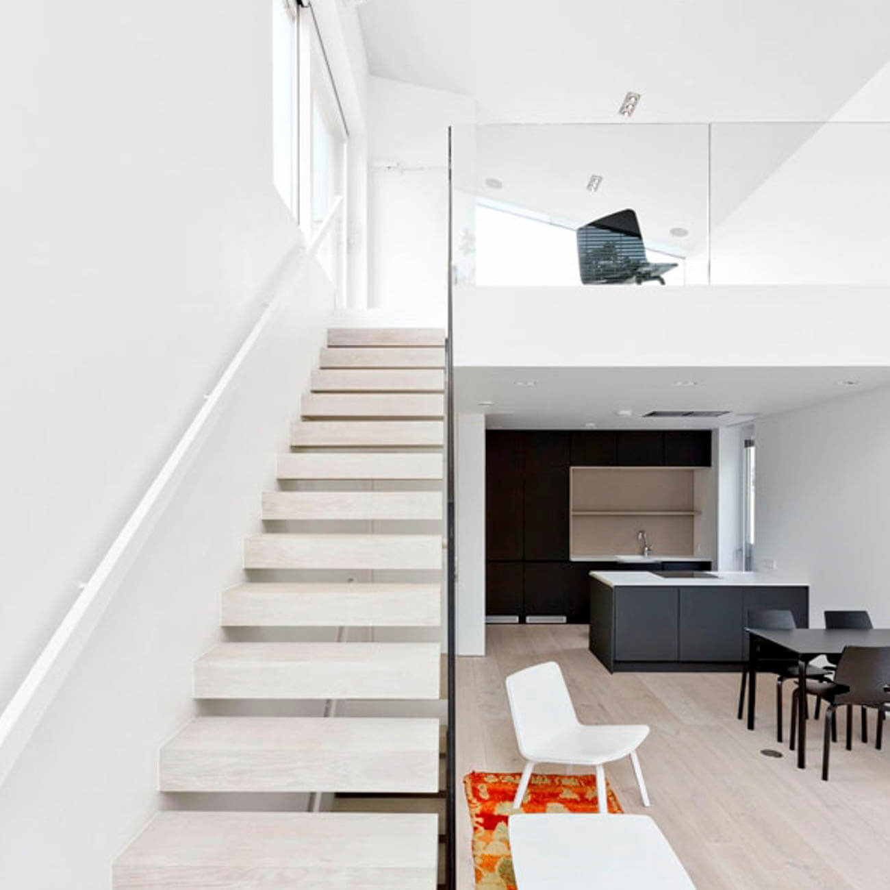 Moderni sisustus valkoisilla portailla, avokeittiö tummilla kaapeilla ja yläkerrassa työtuoli valoisassa tilassa.