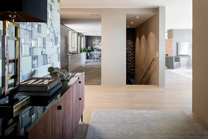 Moderni sisustus, jossa on puinen lipasto, kirja ja koriste-esine, avoin keittiö ja ruokailutila, sekä portaikko ruskealla seinällä ja valkoisella matolla.