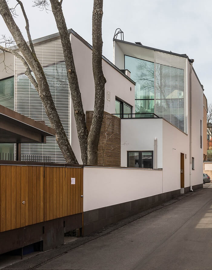 Moderni, valkoinen talo, jossa on suuret ikkunat ja puita edessä, sijaitsee kapealla kadulla. Valoisa sää.