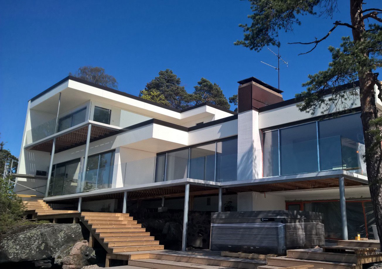 Moderni, valkoinen kaksikerroksinen talo, jossa on suuret ikkunat, lasikaiteet ja puuportaat, aurinkoisessa säässä.