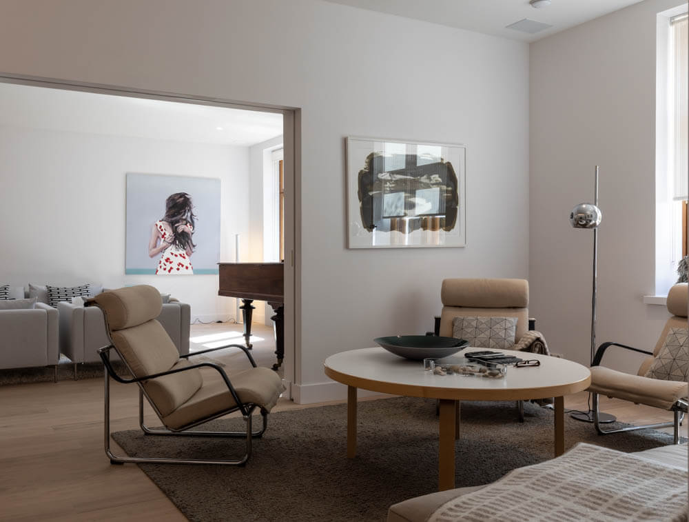 Valoisassa olohuoneessa on beigejä nojatuoleja, pyöreä pöytä, taideteoksia seinillä ja näkymä toiseen huoneeseen, jossa on piano ja sohva.