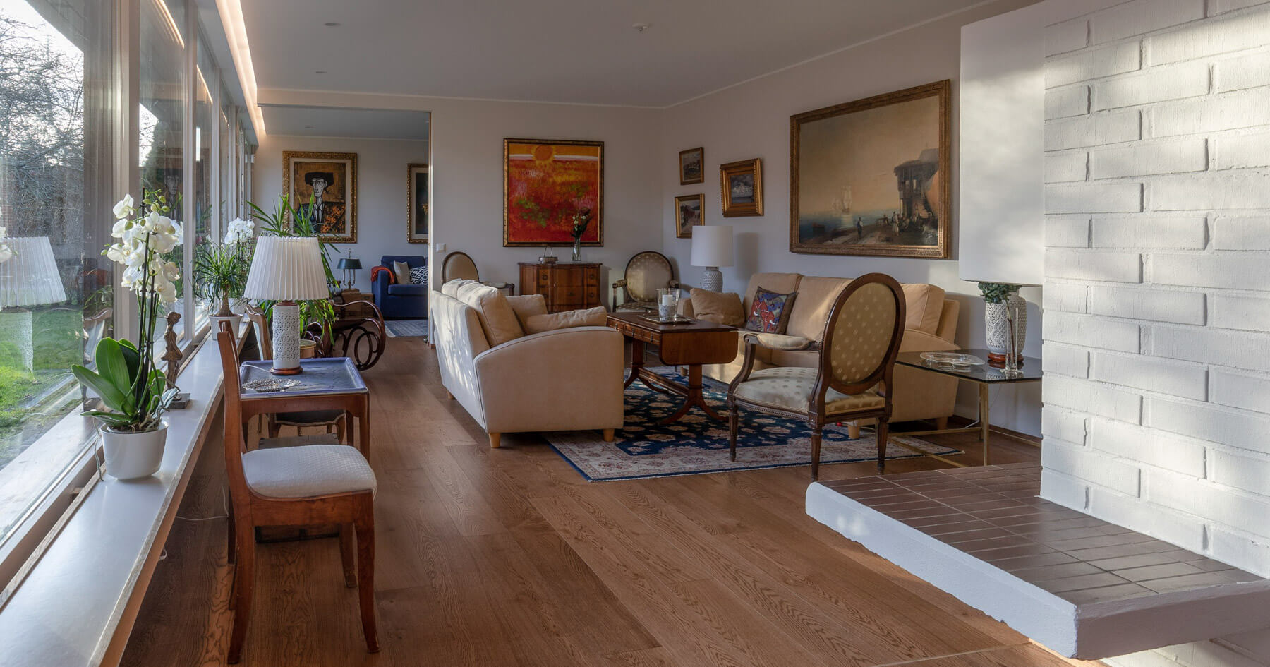 Valoisassa olohuoneessa on suuret ikkunat, vaaleat seinät, puulattia ja valkoinen tiilitakka. Huoneessa on sohvia, tuoleja, tauluja ja kasveja. Aurinko paistaa sisään.