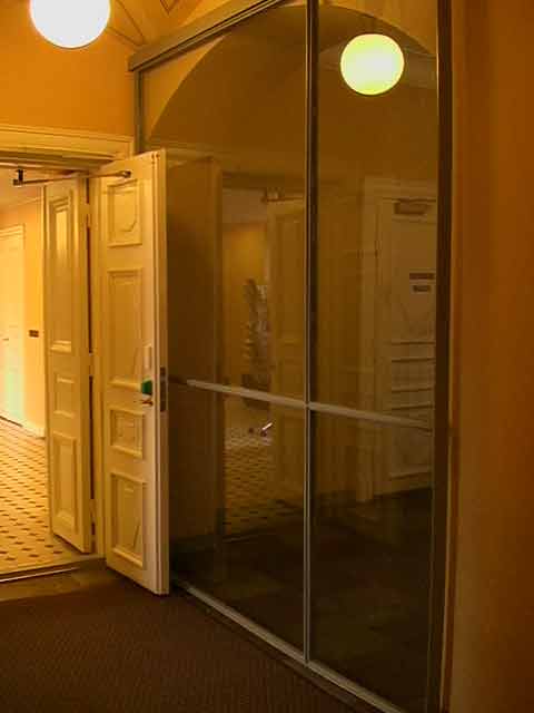 Sisäänkäynti avoimella valkoisella ovella, jossa on vihreä kahva, ja suuri lasiseinä, jossa on heijastuksia ja valaisimia.