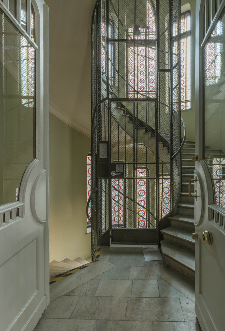 Avoimesta ovesta näkymä vanhaan rakennukseen, jossa on metalliverkolla suojattu kierreportaikko ja värilliset lasi-ikkunat. Lattiat ovat vaaleaa marmoria ja seinät vaaleanvihreät.