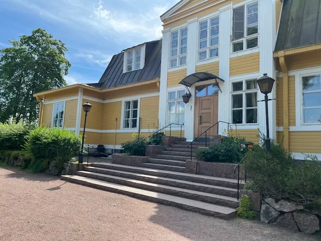 Keltaisen puutalon kiviset portaat, lyhtypylväät ja vihreät pensaat valoisassa säässä.