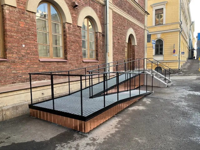 Kuva esittää metallisen pyörätuoliramppi, jossa on mustat kaiteet ja kuparinvärinen pohja, rakennuksen edessä. Rakennus on punatiilinen ja siinä on kaari-ikkunat. Taustalla näkyy keltaisia rakennuksia ja katu. Valo on kirkas ja tasainen.