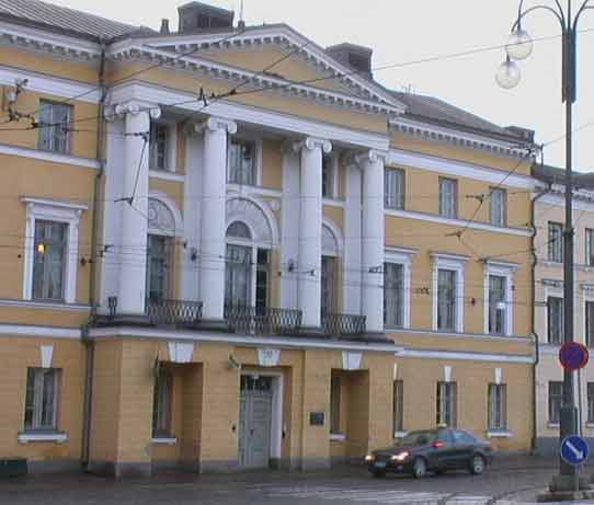 Vaaleankeltainen rakennus, jossa on valkoiset pylväät ja tumma auto edessä, valaistus on päivänvaloa.