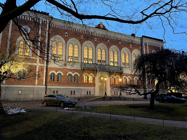 Punatiilinen rakennus, jossa on suuret ikkunat ja koristeellinen julkisivu, valaistu iltavalaistuksessa, etualalla puita ja autoja.