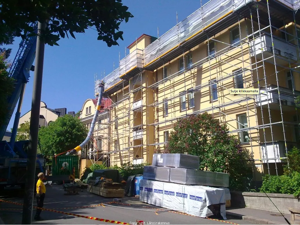 Rakennustyömaa keltaisella rakennuksella, telineet, putki ja materiaaleja kadulla aurinkoisena päivänä.