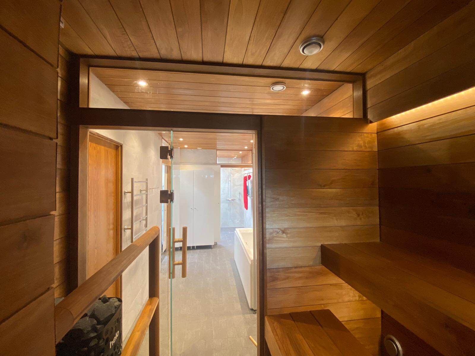 Sauna, jossa puupanelointi, lasiovi ja näkymä kylpyhuoneeseen, jossa on valkoiset kaapit ja kylpyamme.