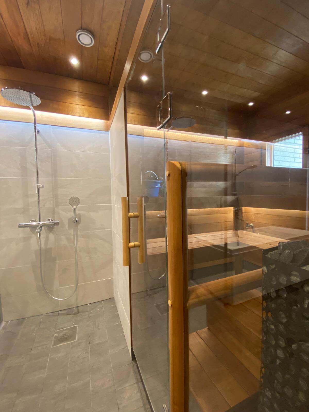 Moderni kylpyhuone, jossa on suihku ja sauna, puupaneloitu katto, harmaat laatat ja lasiseinä.