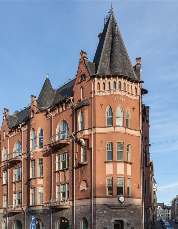 Punatiilinen rakennus, jossa on torni ja koristeellisia yksityiskohtia, valaistu auringonvalossa sinisen taivaan alla.