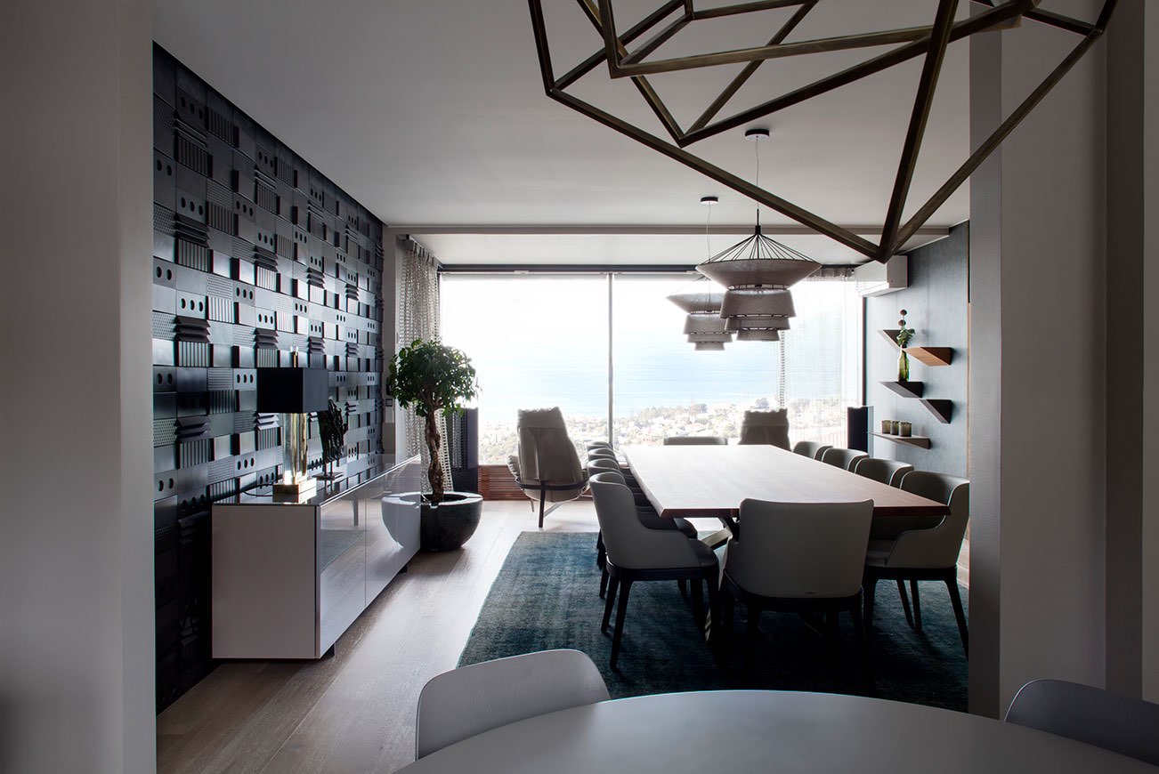 Moderni ruokailutila, jossa on puinen ruokapöytä, harmaat tuolit, koristeellinen seinäpaneeli ja geometrinen kattokruunu. Suuret ikkunat tarjoavat näkymät merelle.