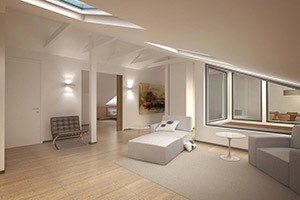 Valoisa ullakkoasunto, jossa on kalteva katto, kattoikkunat, vaaleat puulattiat ja moderni sisustus, mukaan lukien harmaa sohva ja lepotuoli.