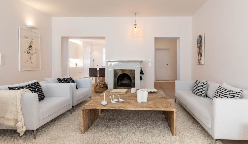 Vaaleassa olohuoneessa on kaksi valkoista sohvaa, puinen sohvapöytä, takka ja taideteoksia seinillä. Huoneessa on pehmeä valaistus ja valkoinen, paksu matto.