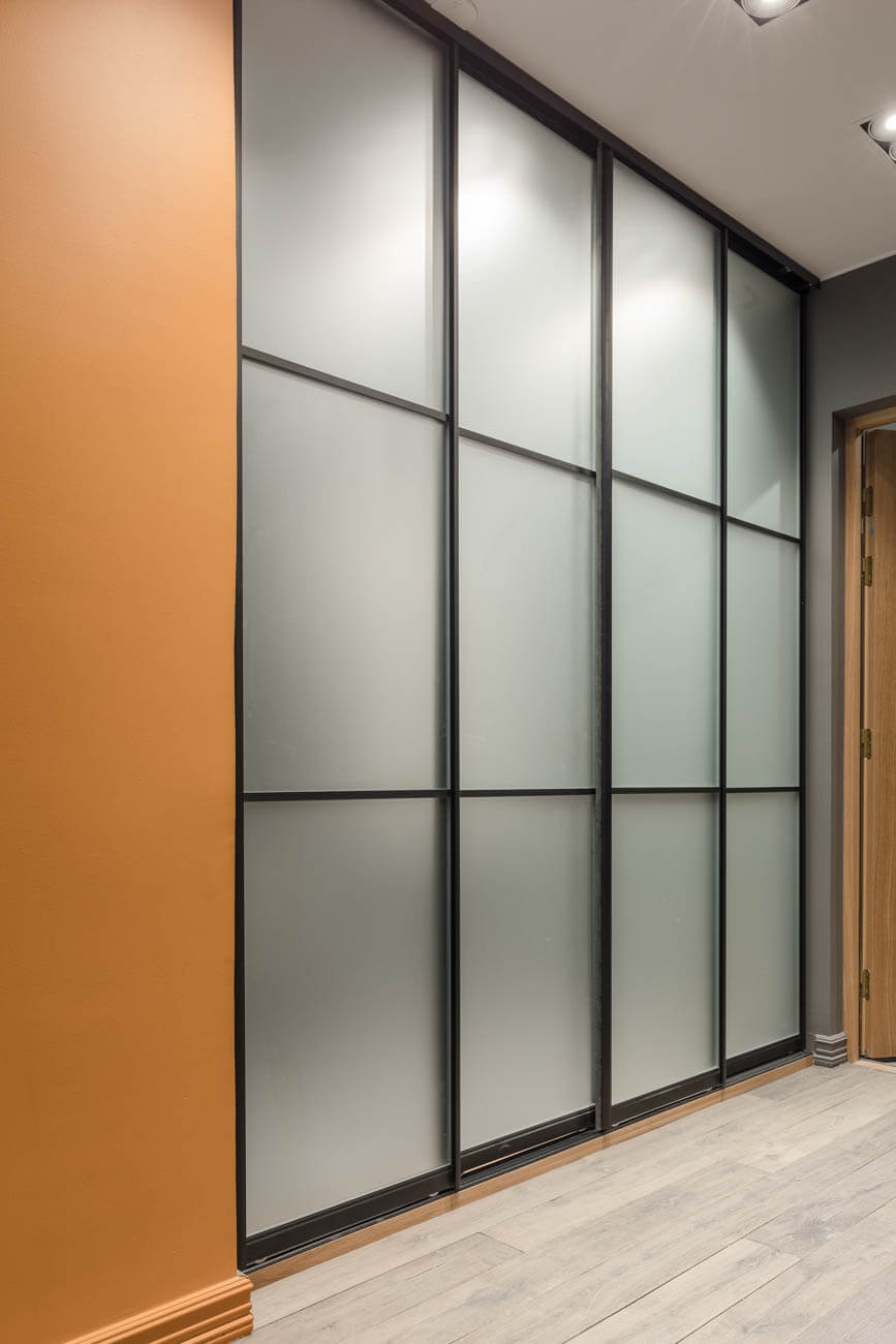 Oranssi seinä, suuri vaatekaappi, jossa on himmeät lasiovet ja mustat kehykset, sekä vaaleat puulattiat.