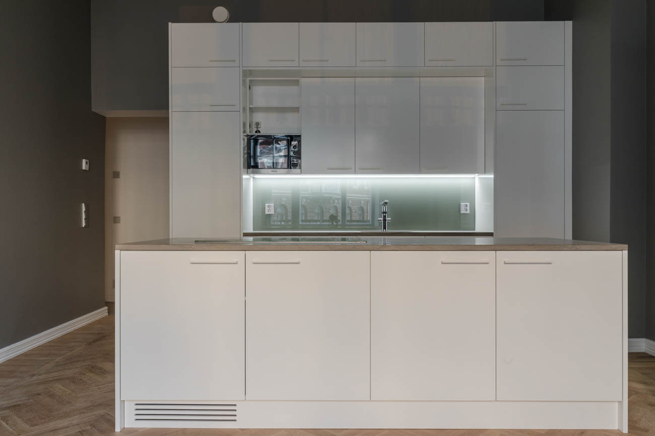 Moderni keittiö, jossa valkoiset kaapit, harmaa työtaso ja parkettilattia. Yläkaapeissa mikroaaltouuni ja valaistu lasinen taustalevy.