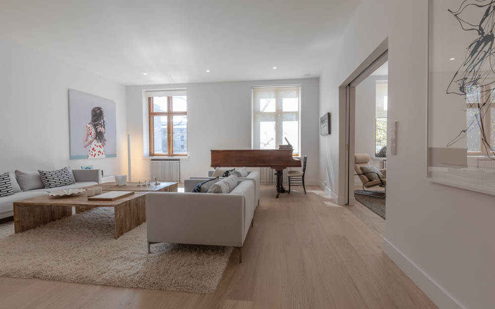 Vaaleassa olohuoneessa on vaalea puulattia, kaksi vaaleaa sohvaa, puinen sohvapöytä, piano ikkunan edessä ja taulu seinällä.