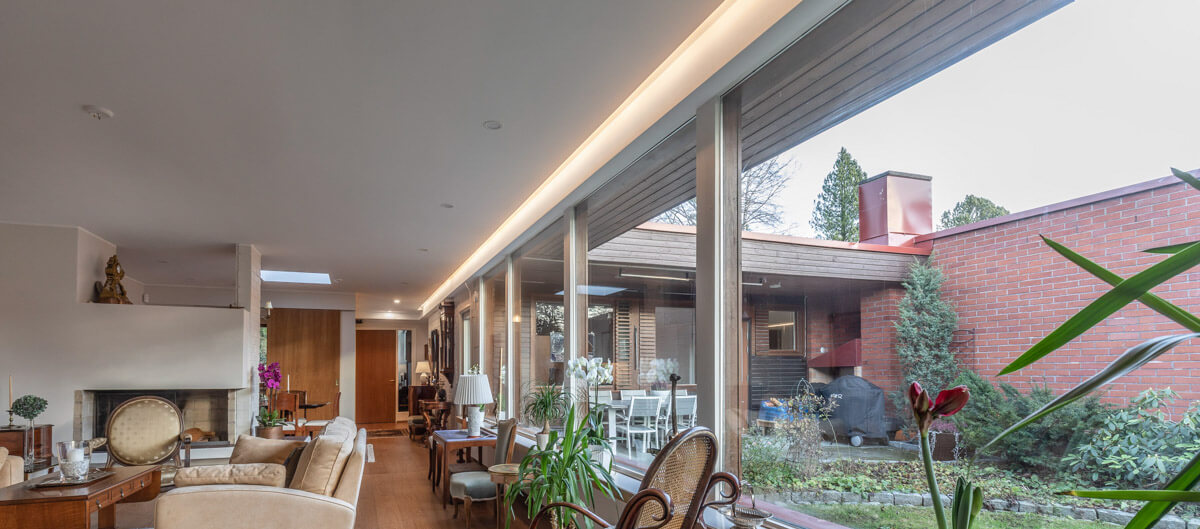 Valoisa olohuone, jossa on suuret ikkunat, puukalusteet ja takka. Ulkona näkyy tiiliseinä ja vihreää kasvillisuutta.